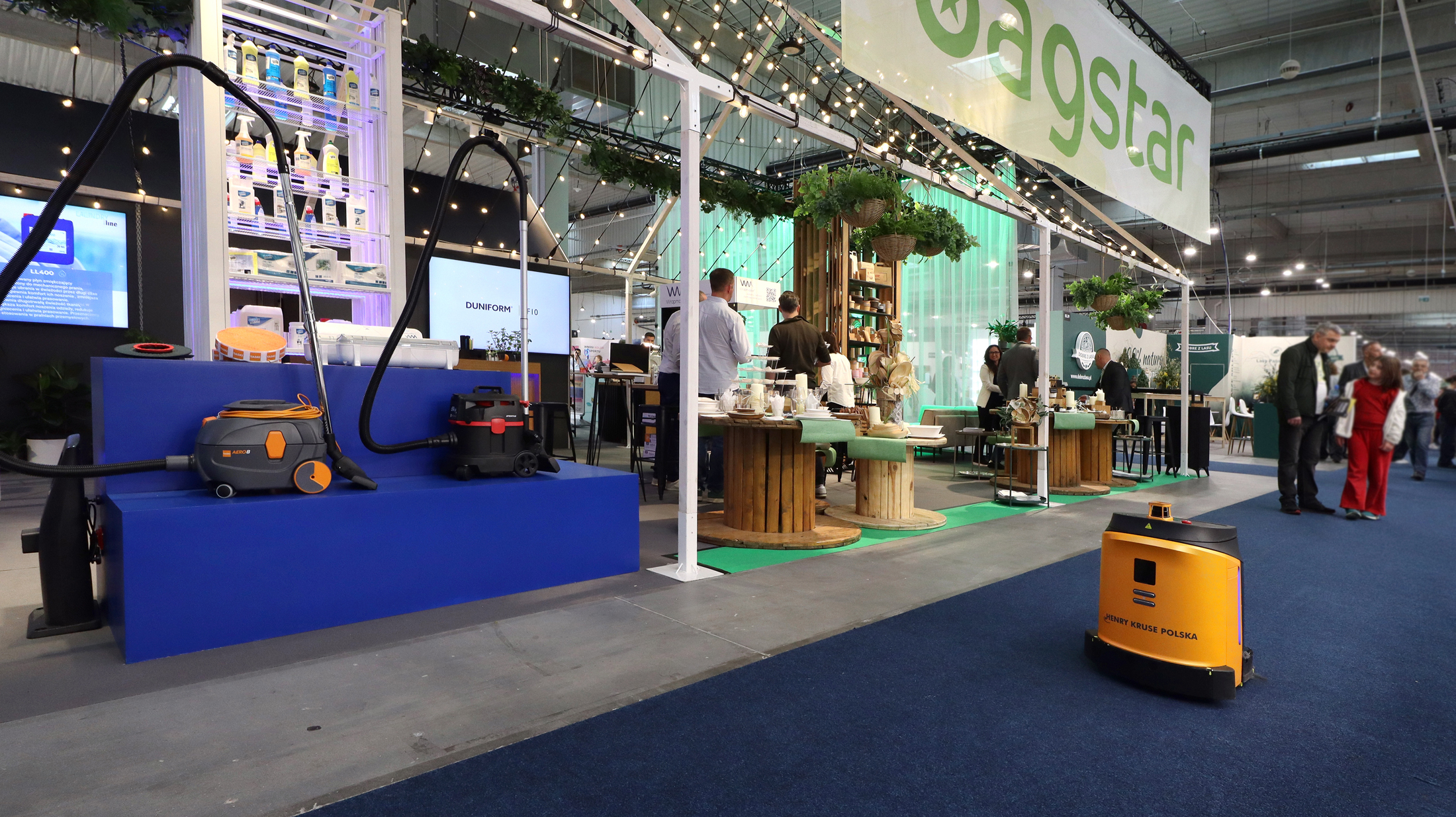 Stoisko targowe prezentuje ekologiczne produkty i rośliny pod znakiem "agstar". Ludzie wchodzą w interakcje przy drewnianych stołach. Z przodu, na niebieskiej wykładzinie widoczny jest sprzęt czyszczący i żółty robot czyszczący podłogę. Otoczenie to kryta hala wystawowa. projekt i nadzór Tatjana Kobuszewska trksystem.com