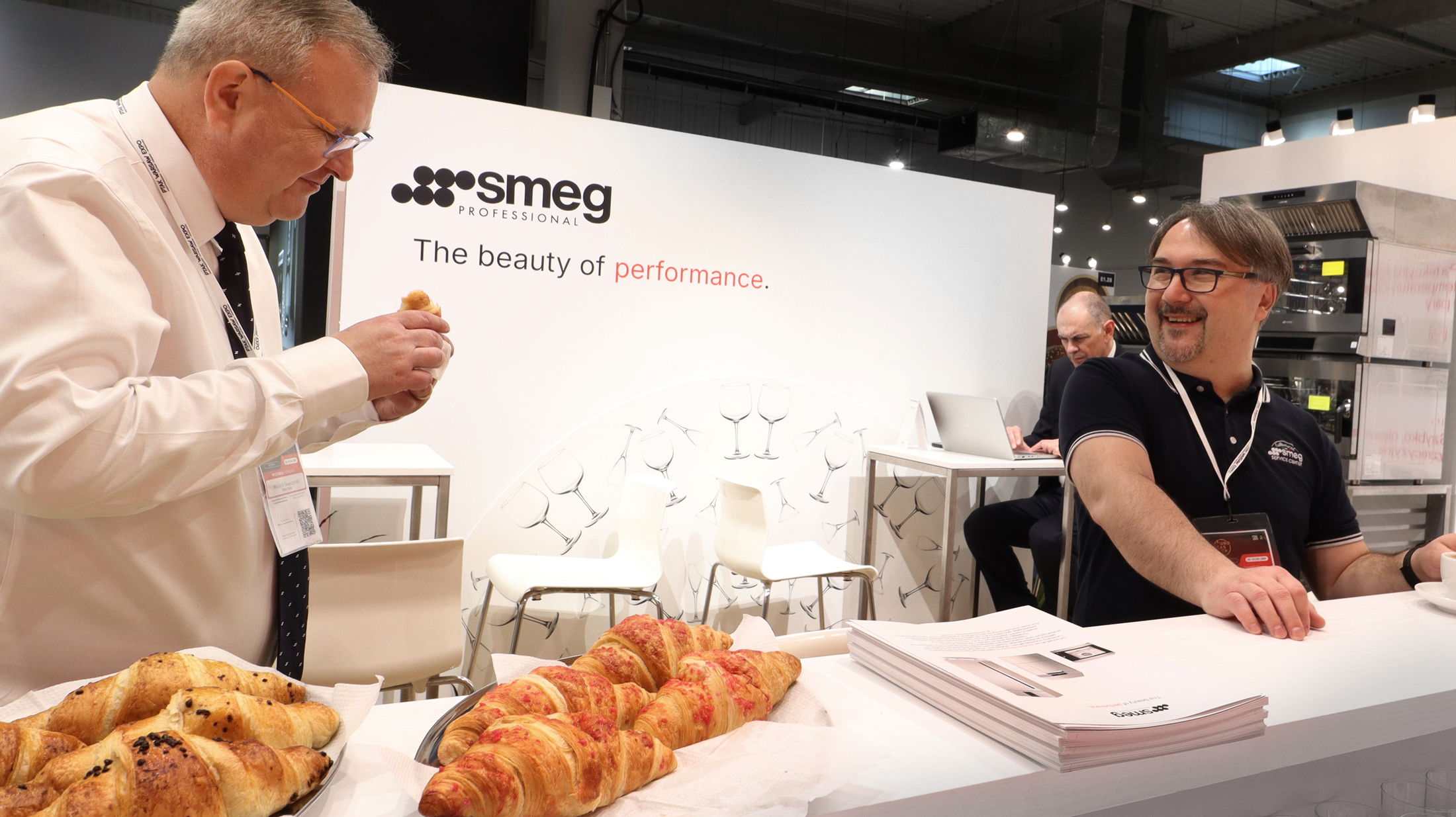 Dwóch mężczyzn wchodzi w interakcję na stoisku targowym z profesjonalnym znakiem Smeg za nimi. Jeden z nich próbuje rogalika, podczas gdy drugi, uśmiechnięty, siedzi za ladą z broszurami i pytaniami i odpowiedziami FaQ. Inna osoba pracuje na laptopie w tle. projekt i nadzór Tatjana Kobuszewska trksystem.com