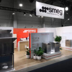 Nowoczesne stoisko targowe dla SMEG Professional obejmuje urządzenia kuchenne ze stali nierdzewnej, rośliny doniczkowe, drewnianą podłogę, czerwono-biały branding i duże logo SMEG powyżej. Stoisko jest dobrze oświetlone w przestronnej hali wystawowej. projekt i nadzór Tatjana Kobuszewska trksystem.com
