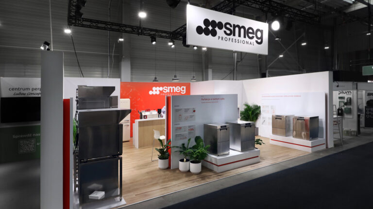 Nowoczesne stoisko targowe dla SMEG Professional obejmuje urządzenia kuchenne ze stali nierdzewnej, rośliny doniczkowe, drewnianą podłogę, czerwono-biały branding i duże logo SMEG powyżej. Stoisko jest dobrze oświetlone w przestronnej hali wystawowej. projekt i nadzór Tatjana Kobuszewska trksystem.com