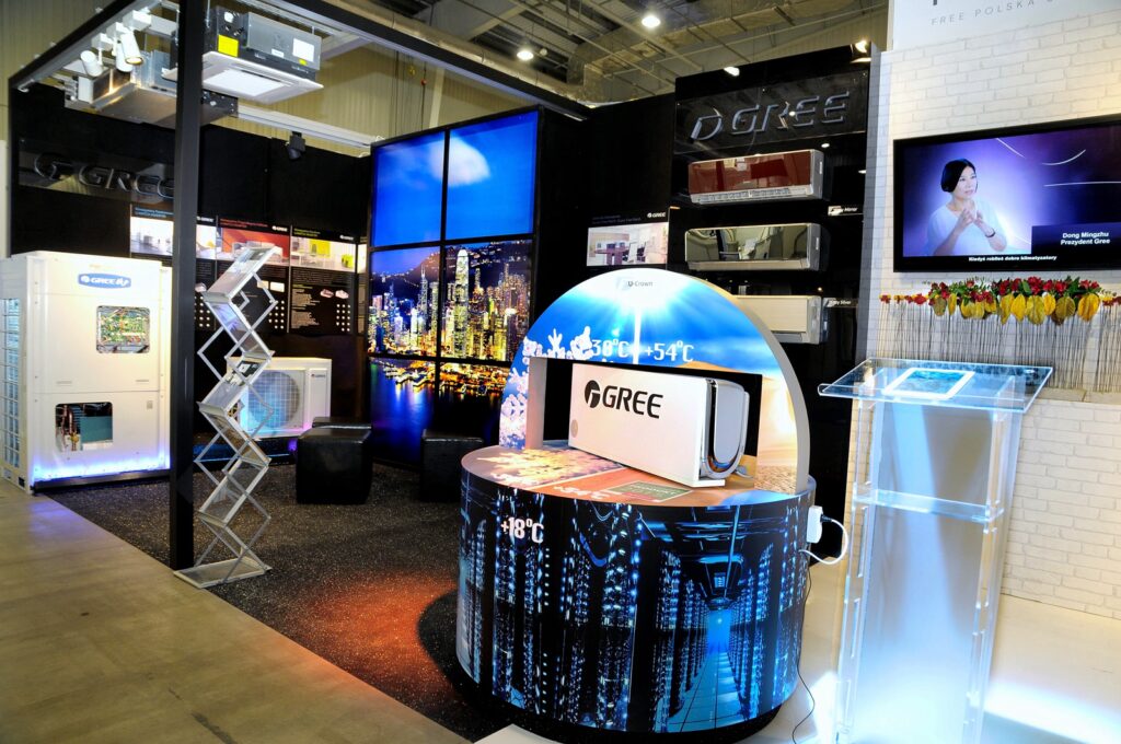 Nowoczesne stoisko targowe na Ptak Warsaw Expo prezentuje urządzenia GREE HVAC z podświetlanymi panelami miejskimi i ekranami cyfrowymi. Okrągły stół z urządzeniem GREE znajduje się w centrum, obok przejrzystego podium i stojaka na broszury po prawej stronie. projekt i nadzór Tatjana Kobuszewska trksystem.com