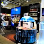 Nowoczesne stoisko targowe na Ptak Warsaw Expo prezentuje urządzenia GREE HVAC z podświetlanymi panelami miejskimi i ekranami cyfrowymi. Okrągły stół z urządzeniem GREE znajduje się w centrum, obok przejrzystego podium i stojaka na broszury po prawej stronie. projekt i nadzór Tatjana Kobuszewska trksystem.com