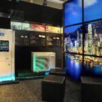 Nowoczesny showroom, zainspirowany Międzynarodowymi Targami HVAC w Ptak Warsaw Expo, obejmuje klimatyzatory Gree, wyświetlacze techniczne na czarnych ścianach i jasną fototapetę z krajobrazem miasta. Trzy czarne fotele w kształcie sześcianu znajdują się na nakrapianej podłodze, tworząc elegancki, nowoczesny klimat. projekt i nadzór Tatjana Kobuszewska trksystem.com