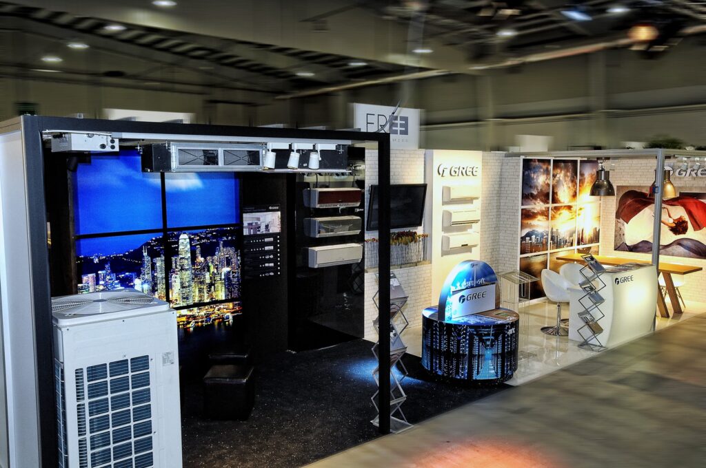 Nowoczesne stoisko wystawiennicze w Ptak Warsaw Expo prezentuje urządzenia HVAC, kanały i systemy wentylacyjne. Banery z napisem "GREE". Oświetlenie dekoracyjne, obrazy miasta i stoiska produktowe wypełniają przestronną, dobrze oświetloną halę. projekt i nadzór Tatjana Kobuszewska trksystem.com