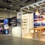 Nowoczesne stoisko wystawiennicze Międzynarodowych Targów HVAC prezentuje urządzenia klimatyzacyjne na białych ścianach z logo "GREE". Drewniany stół i białe krzesła stoją pod wiszącymi lampami, na tle panoramy miasta w Ptak Warsaw Expo. projekt i nadzór Tatjana Kobuszewska trksystem.com