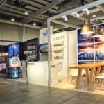 Nowoczesne stoisko wystawiennicze na Międzynarodowych Targach Ptak Warsaw Expo prezentuje klimatyzatory i urządzenia Gree HVAC. Stoisko zawiera grafikę pejzażu miejskiego, drewniany stół, półki na produkty, lampy wiszące i ekrany cyfrowe w przestronnej hali. projekt i nadzór Tatjana Kobuszewska trksystem.com