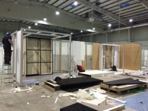 Duża przestrzeń wewnętrzna z wysokimi sufitami przedstawia dwie osoby budujące Stoisko na targi HVAC Ptak Warsaw Expo. Rozrzucone materiały, narzędzia i drewniane palety pokrywają podłogę, podczas gdy niedokończone ściany i ramy są montowane pod jasnym oświetleniem. projekt i nadzór Tatjana Kobuszewska trksystem.com