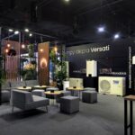 Nowoczesne stoisko wystawiennicze na Międzynarodowych Targach HVAC, Ptak Warsaw Expo, z szarymi fotelami, małymi okrągłymi stolikami, dużymi klimatyzatorami, zielonymi roślinami i wiszącymi lampami. Na czarnych ścianach widnieje polski tekst oraz marki GREE i OLIMPIA SPLENDID. projekt i nadzór Tatjana Kobuszewska trksystem.com