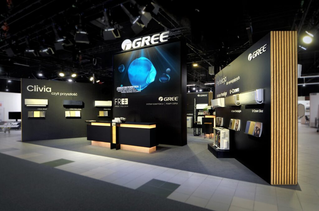 Nowoczesne stoisko targowe GREE z czarnymi ścianami, drewnianymi akcentami i podświetlanymi ladami. Duże ekrany podkreślają produkty HVAC na Międzynarodowych Targach HVAC podczas Ptak Warsaw Expo, z widocznym logo GREE i dobrze oświetlonymi przestrzeniami. projekt i nadzór Tatjana Kobuszewska trksystem.com