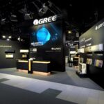 Nowoczesne stoisko targowe GREE z czarnymi ścianami, drewnianymi akcentami i podświetlanymi ladami. Duże ekrany podkreślają produkty HVAC na Międzynarodowych Targach HVAC podczas Ptak Warsaw Expo, z widocznym logo GREE i dobrze oświetlonymi przestrzeniami. projekt i nadzór Tatjana Kobuszewska trksystem.com