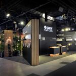 Nowoczesne stoisko targowe dla GREE na Ptak Warsaw Expo ma czarne ściany z zamontowanymi jednostkami HVAC, recepcją i akcentami z drewnianych listew. Rośliny w rogu dodają zieleni, a reflektory oświetlają przestrzeń Międzynarodowych Targów HVAC. projekt i nadzór Tatjana Kobuszewska trksystem.com