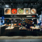 Nowoczesne stoisko wystawowe prezentuje eleganckie nakrycia stołu i naczynia kuchenne. Nad głową, na czarnej ścianie zamontowane są duże zdjęcia wykwintnych potraw z napisami "Fine Dine" i "AMERBOX". Lampy wiszące oświetlają starannie ułożone stoły i talerze. projekt i nadzór Tatjana Kobuszewska trksystem.com