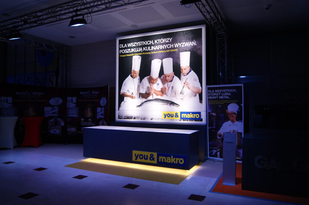 Nowoczesne, słabo oświetlone stoisko koprzedstawia duże zdjęcie czterech szefów kuchni w białych uniformach i czapkach, skupionych na gotowaniu. Logo you makro pojawia się na ladzie, a polski tekst powyżej promuje kulinarne wyzwania i nadchodzące konferencje. projekt i nadzór Tatjana Kobuszewska trksystem.com