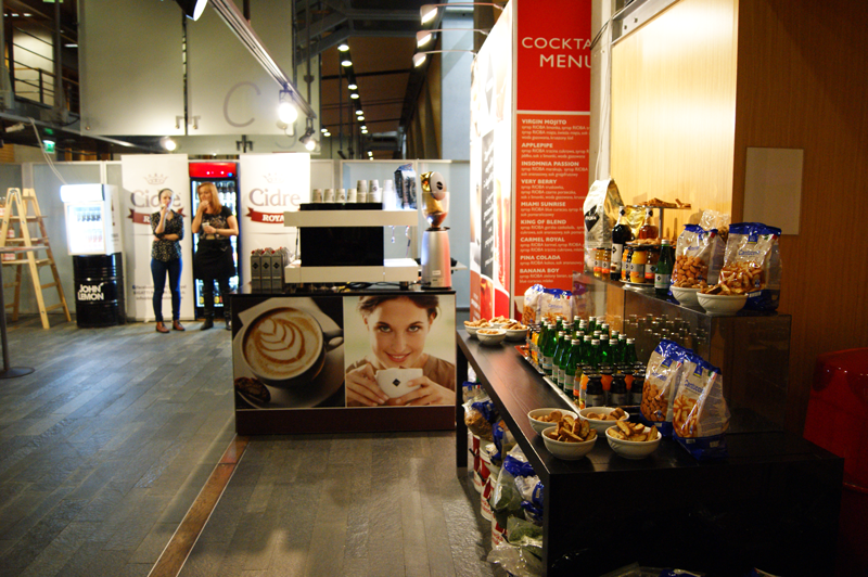 Eleganckie, dobrze oświetlone Stoisko konferencyjne Rioba oferuje nowoczesne ekspresy do kawy, napoje butelkowane oraz przekąski w miseczkach i torebkach. Duży plakat kobiety z filiżanką kawy zdobi ladę, podczas gdy dwie osoby przeglądają ścienne menu z listą koktajli. projekt i nadzór Tatjana Kobuszewska trksystem.com