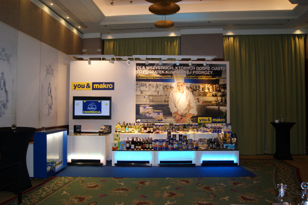 Na konferencji w hotelu, stoisko Makro prezentuje półki z żywnością i napojami pod dużym plakatem przedstawiającym szefa kuchni w kuchni. Wyłożone wykładziną pomieszczenie podkreślają zielone zasłony i dekoracyjne oświetlenie nad głową. projekt i nadzór Tatjana Kobuszewska trksystem.com