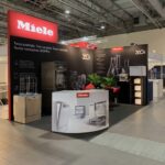 Stoisko targowe Miele zawiera ekspozycje produktów, urządzenia kuchenne i próbki szkła. Czarne ściany wyświetlają logo Miele i polski tekst. Czerwono-białe oznakowanie, lada recepcyjna i rośliny doniczkowe uzupełniają nowoczesną, dobrze oświetloną przestrzeń wystawienniczą. projekt i nadzór Tatjana Kobuszewska trksystem.com
