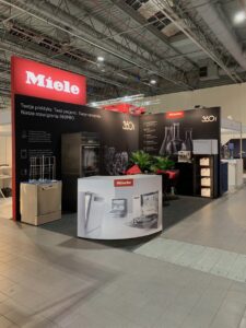 Stoisko targowe Miele zawiera ekspozycje produktów, urządzenia kuchenne i próbki szkła. Czarne ściany wyświetlają logo Miele i polski tekst. Czerwono-białe oznakowanie, lada recepcyjna i rośliny doniczkowe uzupełniają nowoczesną, dobrze oświetloną przestrzeń wystawienniczą. projekt i nadzór Tatjana Kobuszewska trksystem.com