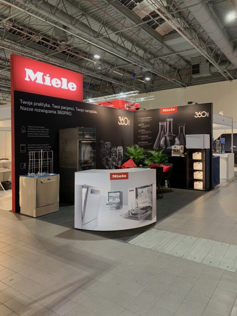 miele stoisko targowe na targach PCI Stoisko targowe Miele zawiera ekspozycje produktów, urządzenia kuchenne i próbki szkła. Czarne ściany wyświetlają logo Miele i polski tekst. Czerwono-białe oznakowanie, lada recepcyjna i rośliny doniczkowe uzupełniają nowoczesną, dobrze oświetloną przestrzeń wystawienniczą. projekt i nadzór Tatjana Kobuszewska trksystem.com