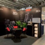 Nowoczesne stoisko targowe z czarnym tłem z logo 360 PRO i Miele, obrazami szklanych naczyń i butelek, małym okrągłym stołem otoczonym czterema czarnymi krzesłami z czerwonymi poduszkami, zielonymi roślinami doniczkowymi i sprzętem czyszczącym z białymi kanistrami. projekt i nadzór Tatjana Kobuszewska trksystem.com