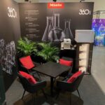 Stoisko targowe z brandingiem "Miele" i "360 Pro" zawiera czarny stół z czterema czarnymi krzesłami, każde z czerwonymi poduszkami. Za stołem znajdują się zielone rośliny doniczkowe, a z boku na czarnych szafkach stoi ekspres do kawy z ustawionymi na sobie mikrofalówkami. projekt i nadzór Tatjana Kobuszewska trksystem.com