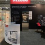 Stoisko targowe Miele z dużym czerwono-białym logo Miele nad polskim tekstem, ze zmywarką na wystawie, broszurami, czarnym dywanem, krzesłami i schematami maszyn w tle pod przemysłowymi lampami sufitowymi. projekt i nadzór Tatjana Kobuszewska trksystem.com