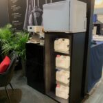 Czarny regał mieści cztery duże białe pojemniki z chemikaliami na przezroczystych półkach. Na górze znajduje się mała biała lodówka i białe urządzenie. Obok znajdują się rośliny, czarna szafka i czarne krzesło. Tło stanowi ciemna ściana ze schematami szkła i tekstem. projekt i nadzór Tatjana Kobuszewska trksystem.com