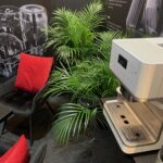 Nowoczesny biały ekspres do kawy stoi na blacie obok bujnych zielonych palm doniczkowych. Obok ustawiono czarne krzesła z jaskrawoczerwonymi poduszkami i szklany stolik. Na ciemnych ścianach widnieją obrazy szklanych naczyń, tworząc przytulny i stylowy kącik wypoczynkowy. projekt i nadzór Tatjana Kobuszewska trksystem.com