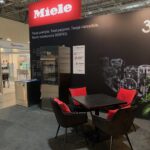 Stoisko targowe z dużym czerwonym logo Miele, czarnymi ścianami i polskim tekstem. Widoczny jest wyświetlacz urządzeń kuchennych, czarny stół z czterema czarnymi krzesłami z czerwonymi poduszkami i jasne oświetlenie górne. W tle widoczne jest inne stoisko. projekt i nadzór Tatjana Kobuszewska trksystem.com