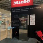 Stoisko wystawowe Miele prezentuje dwie zmywarki do naczyń przed czarną ścianą z dużym czerwonym logo Miele i polskim tekstem. Broszury i stojaki znajdują się na małym stoliku. Stoisko znajduje się w jasnej, nowoczesnej przestrzeni eventowej z innymi stoiskami marki widocznymi w tle. projekt i nadzór Tatjana Kobuszewska trksystem.com