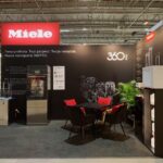 Nowoczesne stoisko targowe z brandingiem Miele, czarnymi ścianami z białym polskim tekstem i zdjęciami produktów 360PRO. Stoisko obejmuje urządzenia, stół z czterema krzesłami, broszury, białą maszynę, rośliny i górne oświetlenie w przestronnej hali. projekt i nadzór Tatjana Kobuszewska trksystem.com