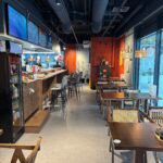 Nowoczesna kawiarnia z drewnianymi stołami, barem po lewej stronie i przytulnym oświetleniem. Wystrój restauracji-halomian-sala-czerwono-czarna obejmuje odsłonięty sufit, jaskrawoczerwoną ścianę akcentującą i duże okna, które wypełniają przestrzeń naturalnym światłem. projekt i nadzór Tatjana Kobuszewska trksystem.com