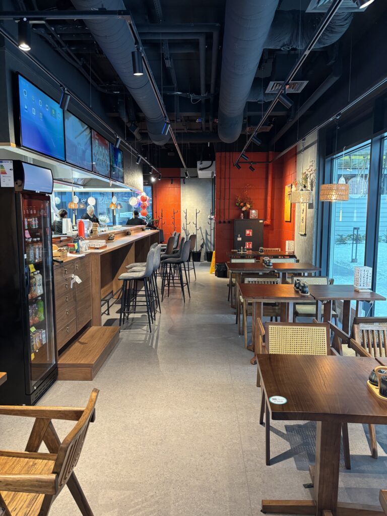 Nowoczesna kawiarnia z drewnianymi stołami, barem po lewej stronie i przytulnym oświetleniem. Wystrój restauracji-halomian-sala-czerwono-czarna obejmuje odsłonięty sufit, jaskrawoczerwoną ścianę akcentującą i duże okna, które wypełniają przestrzeń naturalnym światłem. projekt i nadzór Tatjana Kobuszewska trksystem.com