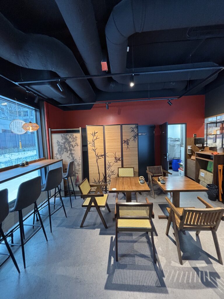 Przytulne wnętrze kawiarni, inspirowane restauracją-halomian-szczegol-wnetrza, obejmuje drewniane krzesła i stoły, bar z wysokimi stołkami przy dużym oknie, bambusowe przegrody, czerwoną ścianę akcentującą i miękkie naturalne światło wpadające do środka. projekt i nadzór Tatjana Kobuszewska trksystem.com