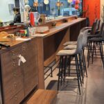 Nowoczesny bar restauracyjny z wysokimi szarymi krzesłami ustawionymi wzdłuż drewnianej lady. Nad barem wiszą lampy wiszące, a czerwoną ścianę w tle zdobią kolorowe lampiony. Osoba stoi za ladą w pobliżu naczyń, szkła i sprzętu kuchennego. projekt i nadzór Tatjana Kobuszewska trksystem.com