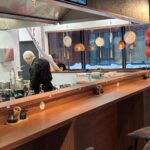 Kucharz z siwymi włosami przygotowuje jedzenie za drewnianą ladą w nowoczesnej azjatyckiej restauracji. Wiszące lampiony i imbryki zdobią przestrzeń. Duże okna odsłaniają śnieg na zewnątrz, a klient siedzi przy ladzie. Ciepłe oświetlenie tworzy przytulną atmosferę. projekt i nadzór Tatjana Kobuszewska trksystem.com