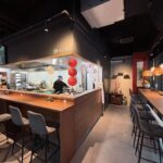 Nowoczesne wnętrze restauracji z drewnianą ladą, szarymi stołkami barowymi i wystrojem w stylu azjatyckim. Szef kuchni pracuje za ladą w pobliżu czerwonych latarni. Na ścianach znajdują się dzieła sztuki i wiszące lampy, tworząc przytulną, współczesną atmosferę. projekt i nadzór Tatjana Kobuszewska trksystem.com