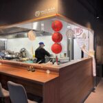 Szef kuchni w czarnym uniformie i niebieskiej czapce przygotowuje jedzenie za drewnianą ladą w Halo Mian, restauracji serwującej makaron. Nad ladą wiszą czerwone chińskie lampiony i ozdobne wachlarze; z przodu stoją trzy szare krzesła. Kuchnia jest czysta i nowoczesna. projekt i nadzór Tatjana Kobuszewska trksystem.com