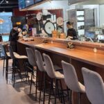 Nowoczesny, czysty bar restauracyjny z drewnianym blatem i szarymi wyściełanymi stołkami. Dwóch pracowników w czarnych uniformach pracuje za ladą, przygotowując jedzenie i napoje. W tle widoczny jest sprzęt kuchenny, waga i bambusowe parowary. projekt i nadzór Tatjana Kobuszewska trksystem.com