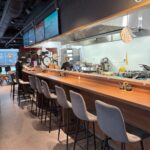 Nowoczesne wnętrze restauracji z długą drewnianą ladą wyłożoną szarymi wyściełanymi stołkami barowymi. Dwie osoby pracują za ladą w otwartej kuchni. Nad głową monitory wyświetlają cyfrowe menu, a latarnie wiszą jako dekoracja. Przestrzeń jest czysta i dobrze oświetlona. projekt i nadzór Tatjana Kobuszewska trksystem.com