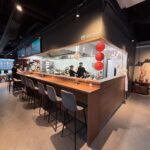 Nowoczesna otwarta kuchnia w restauracji-halomian-bar oferuje kucharzy gotujących za drewnianą ladą wyłożoną szarymi stołkami barowymi. W rogu wiszą czerwone latarnie, a nad nimi świeci napis "HALO MIAN", tworząc ciepłą, zachęcającą atmosferę do swobodnego spożywania posiłków. projekt i nadzór Tatjana Kobuszewska trksystem.com