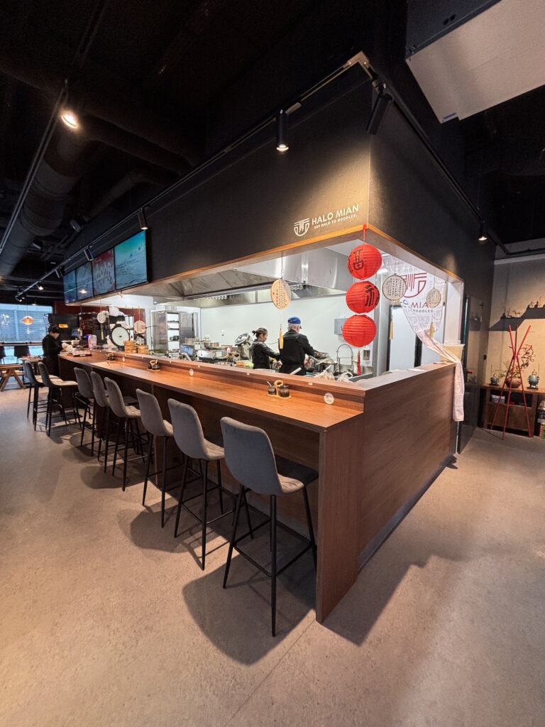 Nowoczesna otwarta kuchnia w restauracji-halomian-bar oferuje kucharzy gotujących za drewnianą ladą wyłożoną szarymi stołkami barowymi. W rogu wiszą czerwone latarnie, a nad nimi świeci napis "HALO MIAN", tworząc ciepłą, zachęcającą atmosferę do swobodnego spożywania posiłków. projekt i nadzór Tatjana Kobuszewska trksystem.com