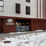 Nowoczesny budynek z brązowo-białą fasadą i dużymi oknami. Na parterze znajduje się restauracja z napisem "HALO MIAN" i "SAY HALO TO NOODLES". Śnieg pokrywa ziemię i chodnik przed wejściem. projekt i nadzór Tatjana Kobuszewska trksystem.com