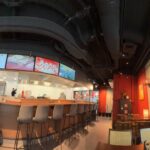 Nowoczesne wnętrze azjatyckiej restauracji z długą drewnianą ladą, wysokimi krzesłami, pracownikiem za ladą, czerwonymi ścianami, cyfrowymi tablicami menu, wiszącymi światłami i oknem odsłaniającym zaśnieżoną miejską scenę na zewnątrz. Stoły i dekoracje są widoczne w całym wnętrzu. projekt i nadzór Tatjana Kobuszewska trksystem.com