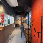 Nowoczesne wnętrze restauracji z czerwoną ceglaną ścianą po prawej stronie, miejscami do siedzenia przy barze obok dużych okien i drewnianą ladą po lewej stronie. Czerwone papierowe lampiony i wiszące światła dodają dekoracyjnych akcentów. Widoczny jest również wieszak na ubrania i rośliny. projekt i nadzór Tatjana Kobuszewska trksystem.com