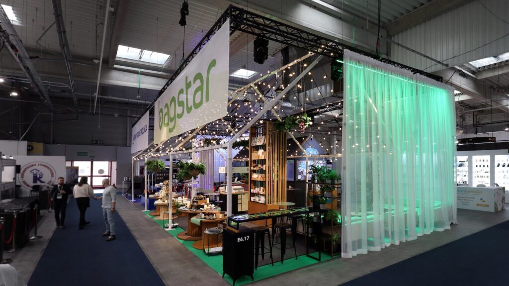 Nowoczesne stoisko targowe Stoisko Bagstar i Henry Kruse na Eurogastro 2026 w Ptak Warsaw Expo zawiera drewniane akcenty, sznurki świetlne, zieleń i starannie ułożone produkty w przestronnej, industrialnej hali wystawienniczej. projekt i nadzór Tatjana Kobuszewska trksystem.com
