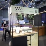 Nowoczesne stoisko targowe prezentuje marki Wrapmaster i bagstar. Na ladach znajdują się białe dozowniki opakowań, artykuły kuchenne i szuflady. Cyfrowy ekran pokazuje żywność w pojemniku. Przestrzeń jest oświetlona górnym światłem, a w tle kręcą się ludzie. projekt i nadzór Tatjana Kobuszewska trksystem.com