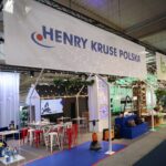 Stoisko targowe z pogrubionymi literami HENRY KRUSE POLSKA nad głową, lampkami sznurkowymi powyżej, wyświetlaczami produktów, stołami i stołkami. W tle widoczny jest zielony znak bagstar i różne urządzenia czyszczące. Otoczenie jest jasne i przestronne. projekt i nadzór Tatjana Kobuszewska trksystem.com