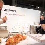W stoisku-smeg-professional-eurogastro- dwóch mężczyzn wchodzi w interakcję, próbując croissanta z tacy z ciastkami. Siedzący mężczyzna uśmiecha się, podczas gdy broszury są ułożone na ladzie, a trzeci mężczyzna pracuje przy laptopie w tle. projekt i nadzór Tatjana Kobuszewska trksystem.com