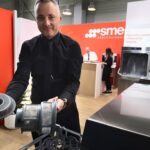 Mężczyzna w czarnej koszuli demonstruje kosz z filtrem do zmywarki w salonie wystawowym. Trzyma filtr nad otwartym stojakiem zmywarki. Za nim dwie osoby rozmawiają przy ladzie z pomarańczowym logo SMEG na ścianie. Rośliny doniczkowe zdobią nowoczesną przestrzeń. projekt i nadzór Tatjana Kobuszewska trksystem.com