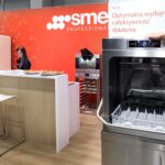 Mężczyzna demonstruje komercyjną zmywarkę do naczyń przed czerwonym stoiskiem SME Professional. Kobieta za drewnianą ladą przygotowuje napoje, a na ladzie stoją białe stołki i jedzenie. Polski tekst na zmywarce tłumaczy się jako Optymalna wydajność i wydajność. projekt i nadzór Tatjana Kobuszewska trksystem.com